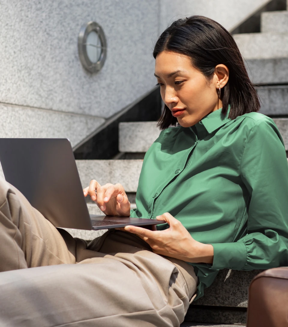 Woman sitting on outdoor steps, working on a laptop and holding a smartphone, focused on her work.