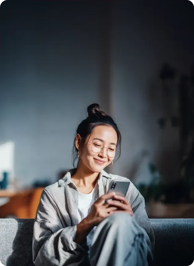 Young woman smiling while using her smartphone, seated indoors with soft natural light and a blurred background.