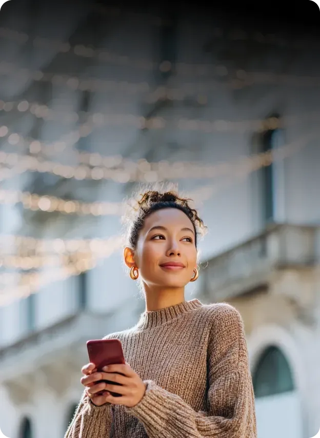 Young woman holding a smartphone outdoors, looking ahead with a smile against a softly lit urban background.