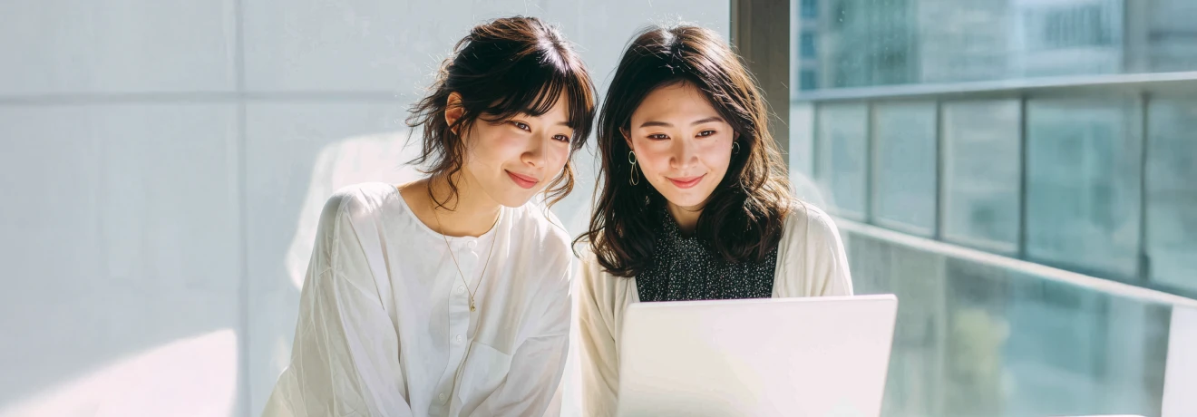 Two women sitting side by side and smiling as they look at a laptop screen in a sunlit modern office with glass windows.
